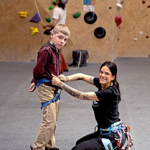 A staff member assists a young participant with his harness in preparation for a rock climbing event. The indoor rock wall is seen in the background. 