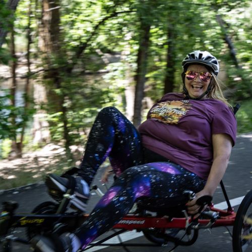 A participant leans back on her recumbent bike and beams as she speeds through a shaded trail.