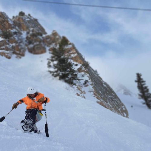 A participant in bright orange winter gear gleefully slides down a steep, snowy slope.
