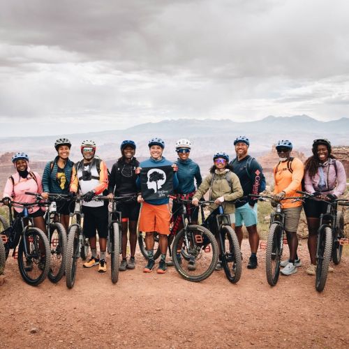 Volunteer Leaders participate in the Capstone experience held in Moab, Utah. They all stand in a line, holding mountain bikes on red clay ground.