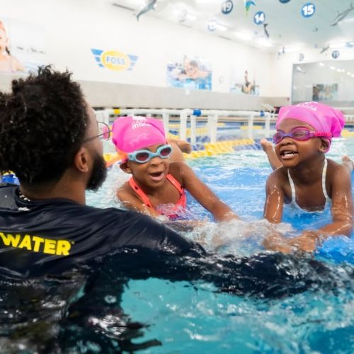 An Outdoor Afro leader guides two young participants with pink swim caps through the water in an indoor pool as they joyfully learn how to swim.