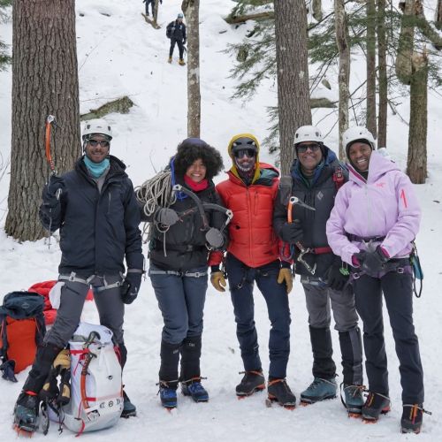 Five participants in cold weather and ice climbing gear take a picture in front of a snow-covered hill to celebrate their winter adventure.