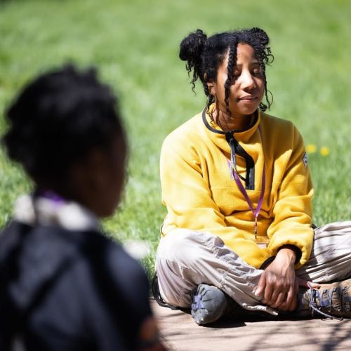 A participant in a yellow sweatshirt sits cross-legged in a grassy area and closes her eyes in concentration during a mental wellness moment.