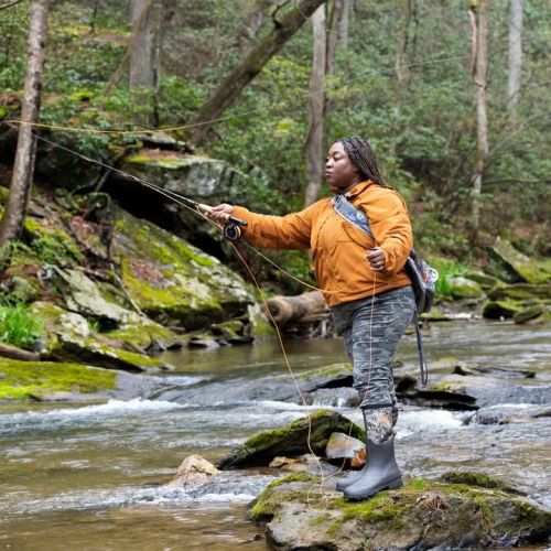 An Outdoor Afro participant stands on a mossy rock in the middle of a river and casts a fishing line. Surrounding the river bank are more mossy rocks and forest undergrowth.