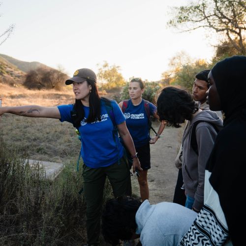 A leader wearing a blue shirt and a backpack points at a sign. Young people stand around looking at the sign among a dry, brush landscape.