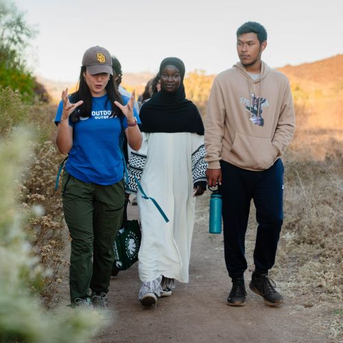 A diverse group walks a path through dry brush. A leader in a blue shirt talks to a girl in a hijab and a boy in a hoodie.