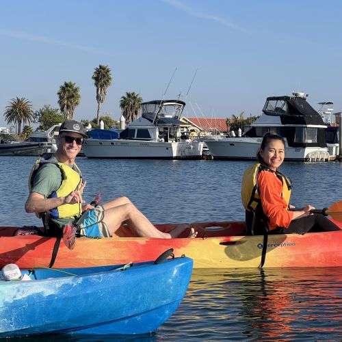 A boy and a girl ride in a tandem kayak. Both hold paddles and wear life vests. The water is calm and there are boats and trees in the background.