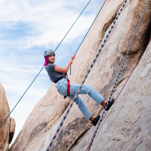 A young girl with pink hair rappels down a rock face using rope. The girl is wearing blue jeans and a helmet.