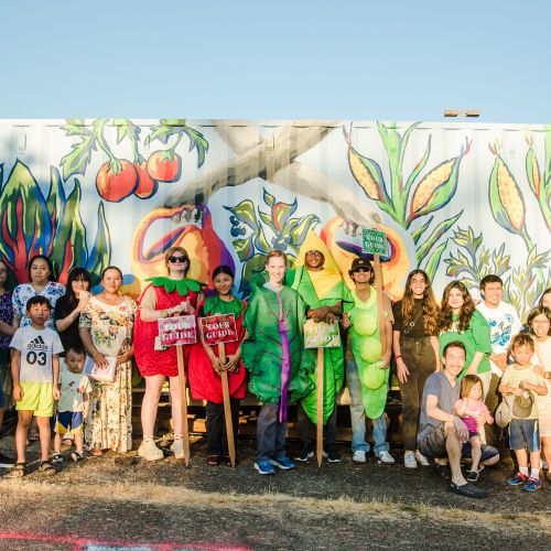 Group of adults and children, some wearing vegetable costumes, standing in a row in front of a colorful storage container with fruits and vegetables painted on it.