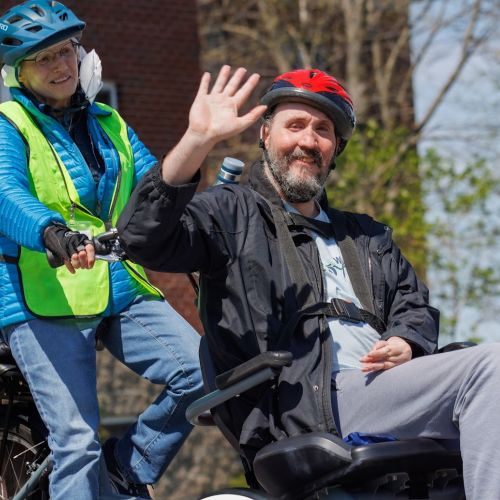 A participant smiles and waves at the camera as he passes by on his trike, with his volunteer looking ahead as she takes him along.