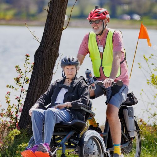 A program staff takes an elderly participant on a bike ride beside a lake. The staff is wearing a neon vest and the bike has an orange flag sticking up in the back. 