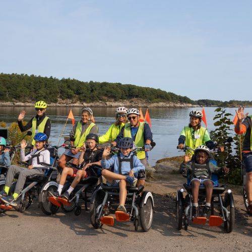 Participants from Peaks Elementary pose in their trikes with their volunteers. Volunteers all wear neon vests. A vast lake, bordered by dense trees, stretches behind them.