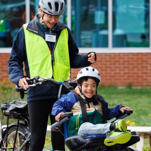 A young participant shows excitement for the bike ride; he is accompanied by a program staff, who smiles down at him. They are riding in front of a brick building with windows. 