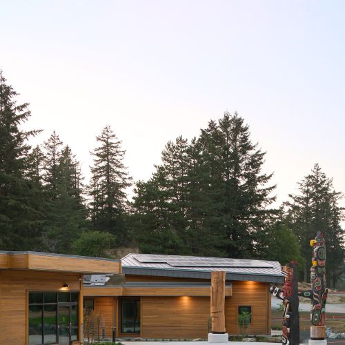 A wood-sided building with a curving walkway, sloped grassy areas, and totem poles.