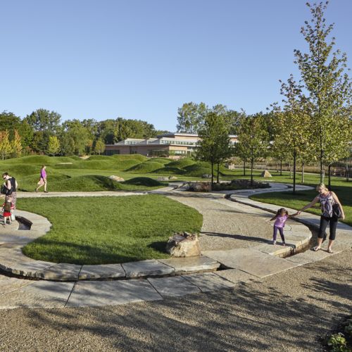 Two children accompanied by adults wander around the curved paths of a water runnel. Another adult is in the background as are mounds of green grass.  