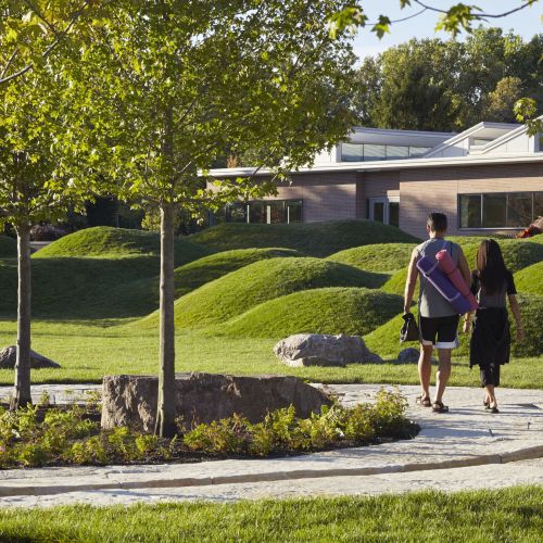 Two people walk on a winding stone path that incorporates a water-filled channel. There are trees on the side of the path, with grass-covered mounds and a building in the background.