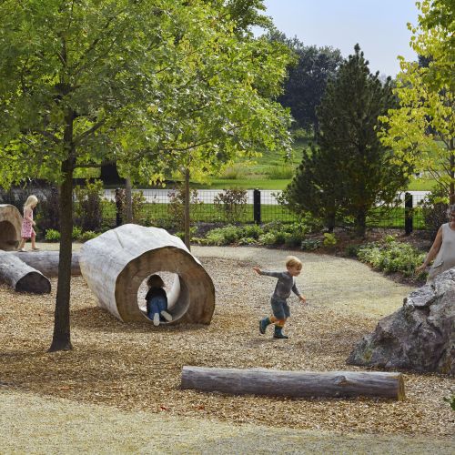 Large mulched area features logs and boulders with children playing while an adult watches.  