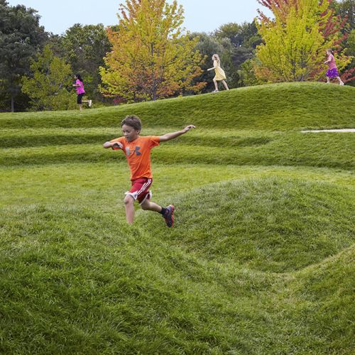 Children run and play on a large expanse of mown grass with irregular mounds and a gentle hillside and trees in the background.