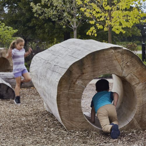 Children play in and around large, hollowed-out logs set in a flat area covered with bark mulch with trees in the background.