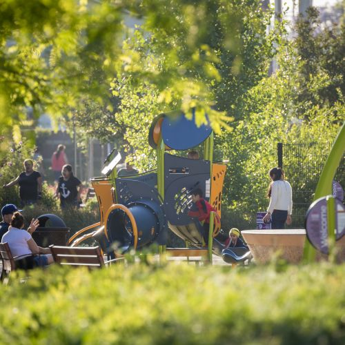 Children play on structures while adults sit on a park bench. There’s greenery in the foreground and background.