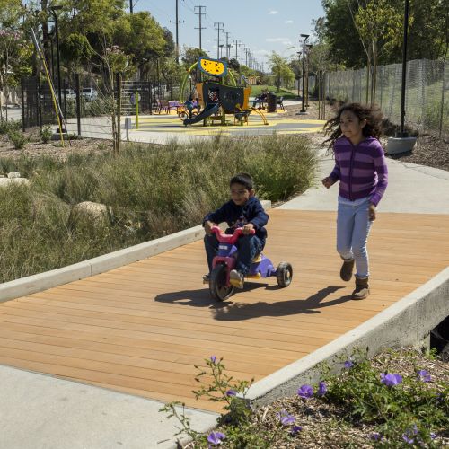 Two children travel along a wide path, one on a tricycle and one running, with a children’s play structure in the background.