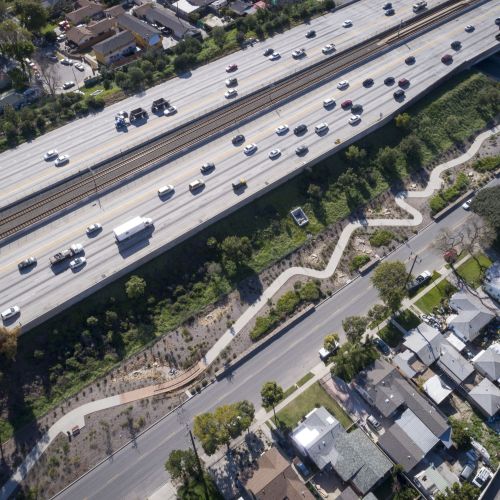 Aerial view shows an elevated freeway in the top left, with a park area and winding path running alongside it. Neighborhood houses fill the rest of the photo.