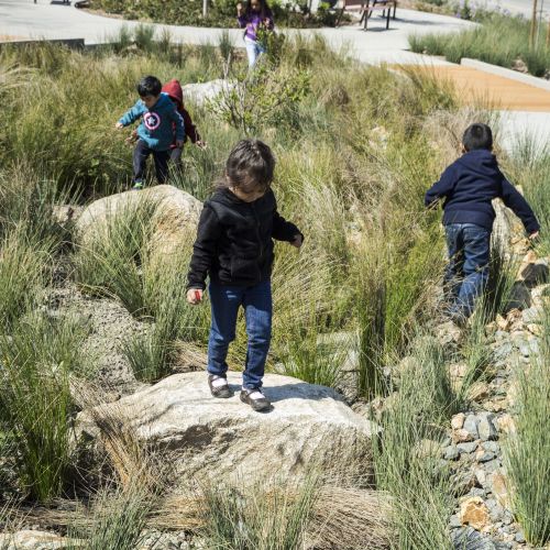 Children walk on and between large boulders in an area with small rocks, gravel, and plants, with a winding path in the background.