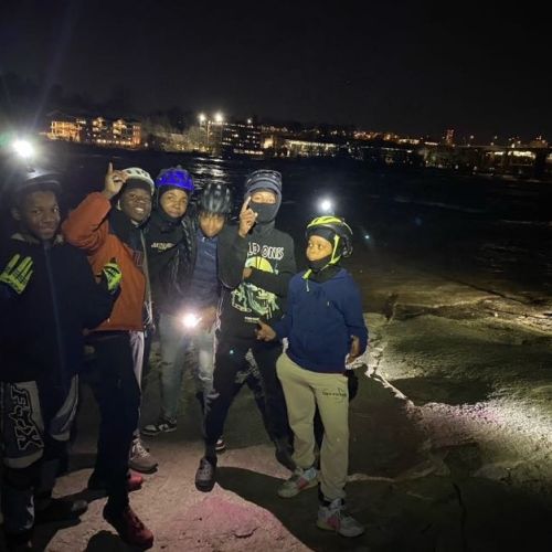 A group of kids in biking helmets happily pose for a nighttime picture. Three hold up their hands to pose while the remaining two remain simply standing. They have flashlights as well as lights attached to their helmets, which illuminate the dark space around them.