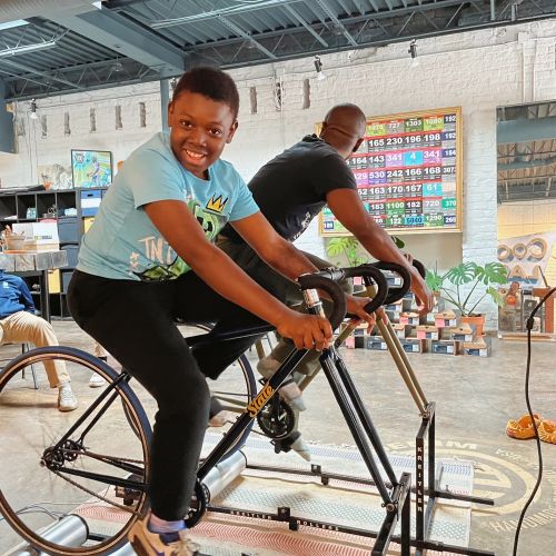 A youth practices his biking skills on an indoor biking machine and beams at the camera. Another person is training on the machine beside the youth, though he is looking away toward the biking equipment lined up in the background.