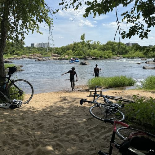Two cyclists leave their bikes behind on the sand shaded by trees. One cyclist is catching up to the other, who is standing at the edge of a lake. The water surface sparkles under the bright, sunny sky, and on the other side of the lake are endless verdant trees that stretch far into the horizon. 