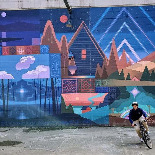 A cyclist cruises along a concrete wall on his bike. On the concrete wall itself is a large mural, which involves various paintings of different scenery that all combine to make the overall mural. The painting includes art of clouds, a house among hills, a geometric depiction of a landscape, and silhouettes of trees that open to reveal a nighttime sky.