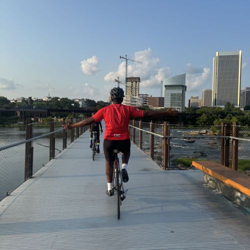A cyclist is cruising along a pier that extends into a lake. Their confidence in biking clearly shows from the way they stretch their arms out instead of holding onto their bike handlebars. The sky overhead is bright blue, with large clouds looming in the distance. On one side of the horizon are large skyscrapers, which tower over the line of trees on the left side of the picture.