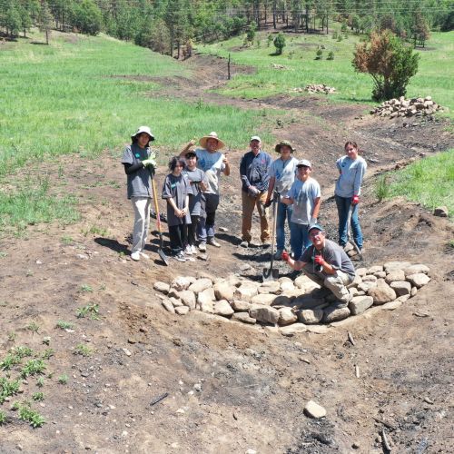 Adults and youth stand by a rock dam built inside a dirt path with grass on the sides and trees in the background.