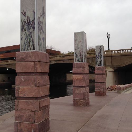 Three stone pillars rise from the edge of a walkway, each one featuring different botanically themed artwork on the top. There’s a bridge in the background and a river on the left.