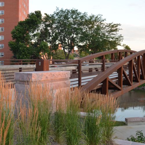 A bridge spans a waterway, with gardens planted with grasses and shrubs in the foreground and trees and a large building in the background.