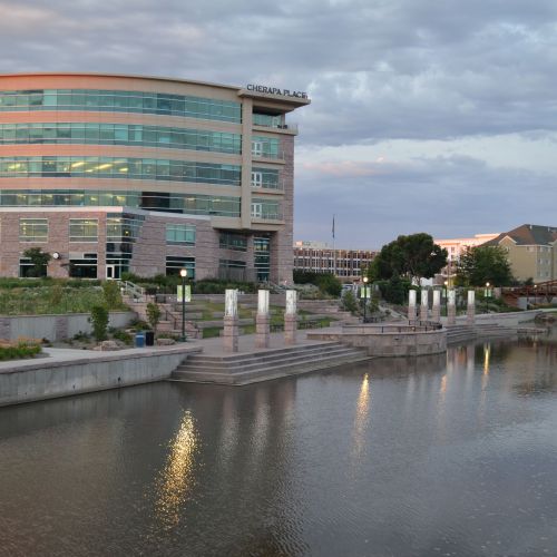 A river is in the foreground. On the riverbank there are steps down to the river, gardens, and grassy areas. There’s a large building in the background.