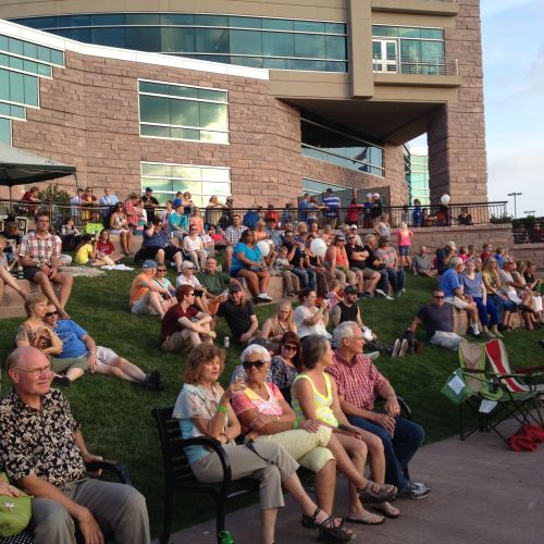A crowd of dozens of people sit on benches and on a grassy slope, most of them facing the same direction, with a large building in the background.