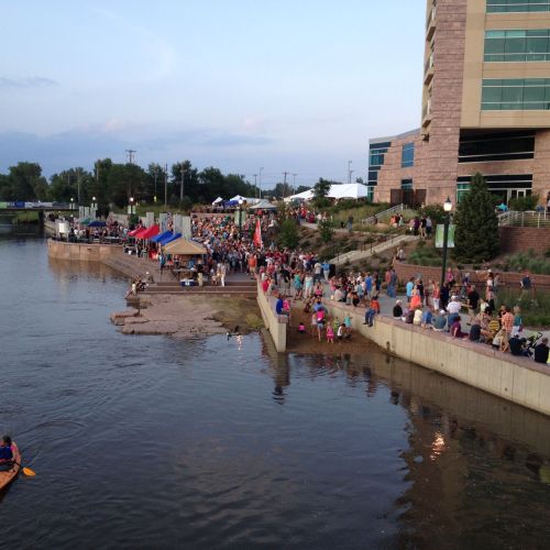 A kayaker paddles in the river. On the riverbank, crowds of people walk on a wide path and sit on a low wall. There’s a large building in the background.