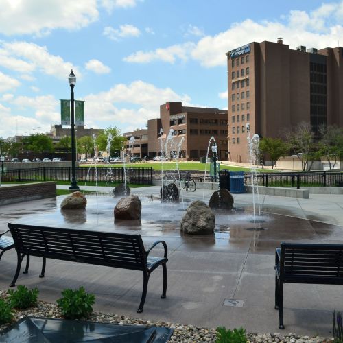 A circular plaza with several benches along the perimeter. Vertical streams of water emerge from the water feature in the center.