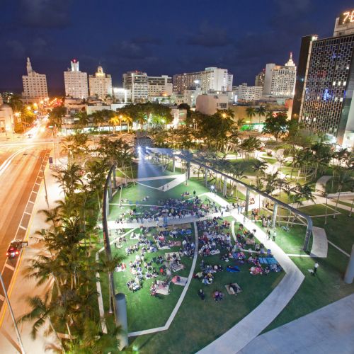 Nighttime aerial view shows the illuminated park with the projector on, a crowd of people sitting on the lawn, and the city skyline in the background.