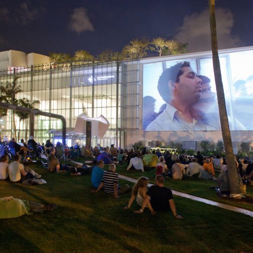 A crowd sits on a grassy area facing toward a large screen on the side of a building. A concert is projected onto the screen.