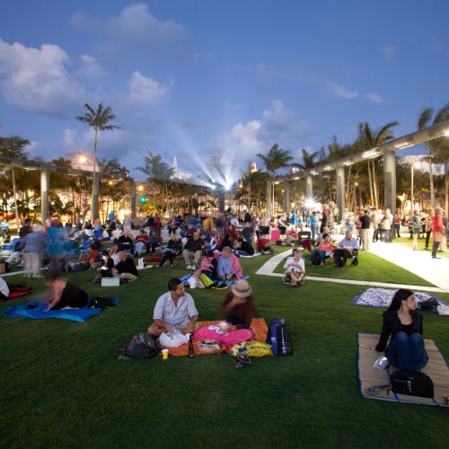A crowd sits on a grassy area at dusk with the projector lights showing in the background. Many people are sitting on blankets.