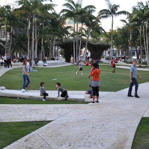 Children and adults stand, walk, and play on wide paths with palm trees in the background.