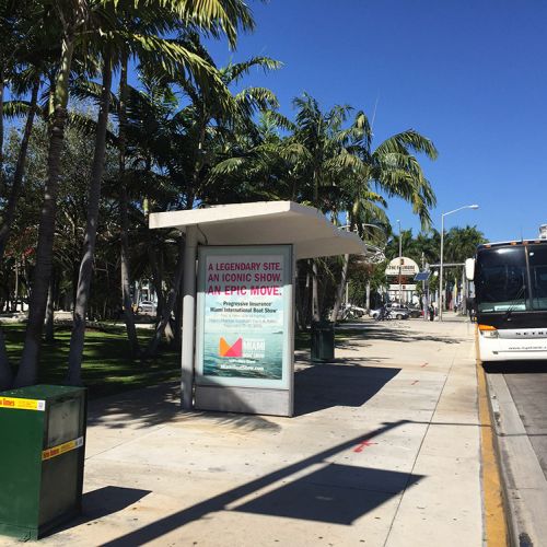Wide sidewalk features a bus stop with palm trees on the left and a road on the right. 