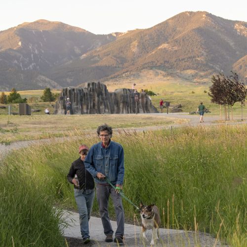 Two people walk a leashed dog on a path. There’s tall grass on either side of the path and mountains in the background.