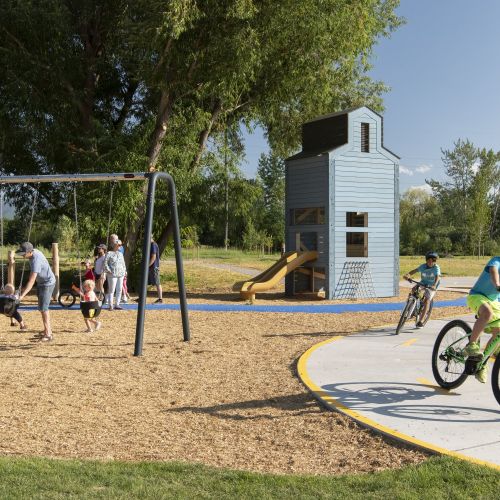 Two children on bicycles on a curved path and several children on swings in a play area. A wooden play tower with slide is in the background next to trees.