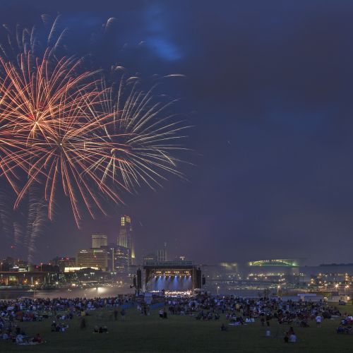 Crowd gathered on a lawn with fireworks in the night sky and cityscape in the background.