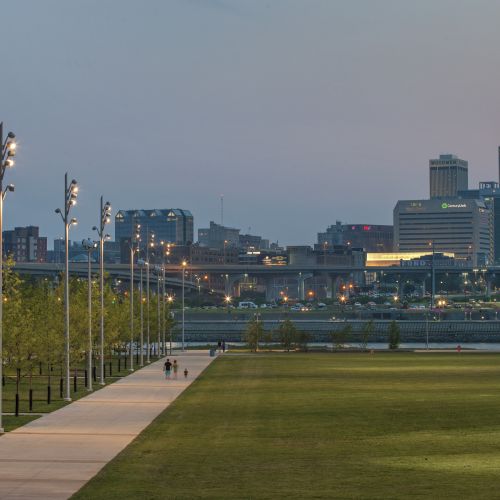 Wide concrete pathway next to a large grassy area with cityscape in the background.