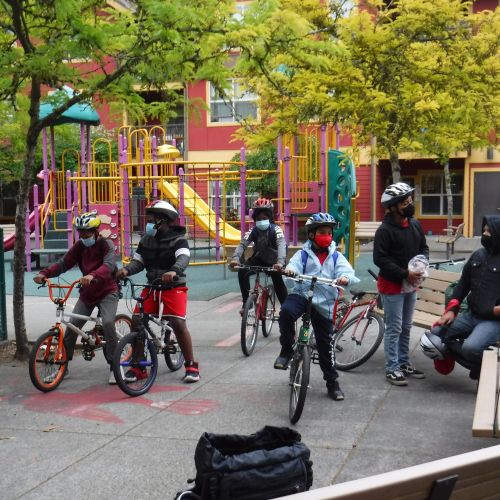 A group prepares for a family friendly biking event during COVID. Behind them is a colorful playground. 