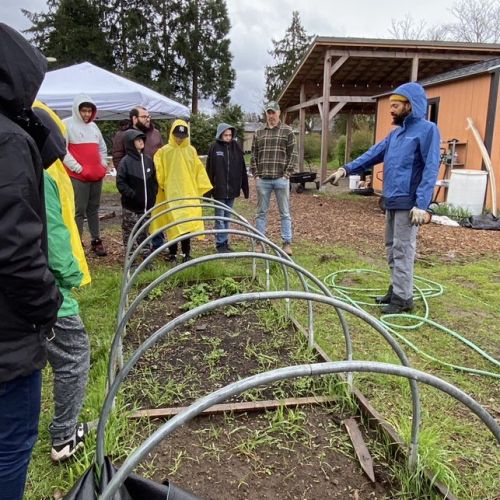 Participants gather around a garden bed in rain gear while an instructor points to the bed. 